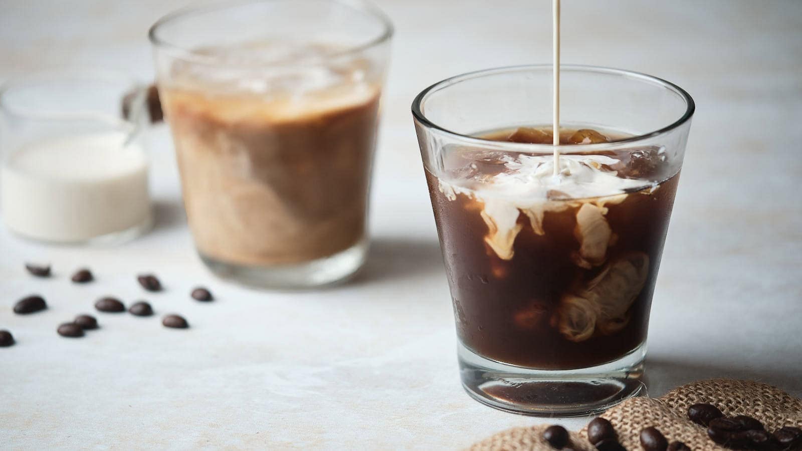 Two glasses of cold brew coffee with milk being poured into one.