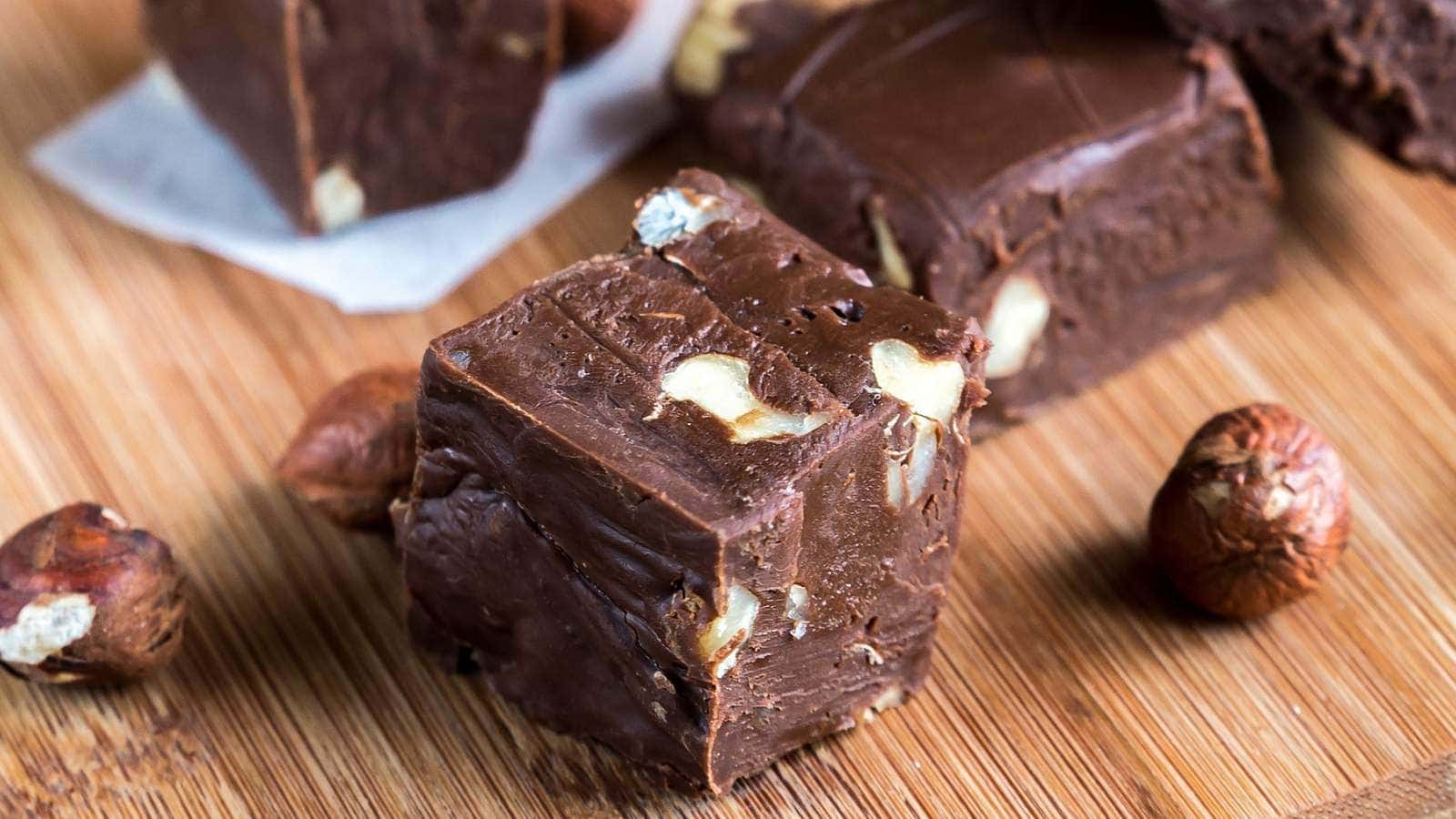 Close-up of nutella fudge squares with nuts on a wooden cutting board, surrounded by whole hazelnuts.