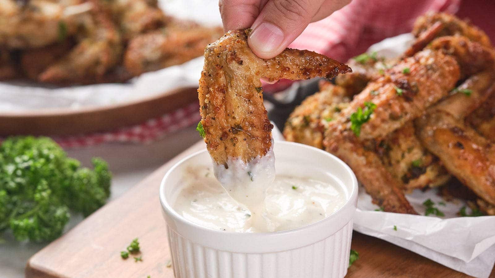 A hand dips a parmesan chicken wing into a white dipping sauce.