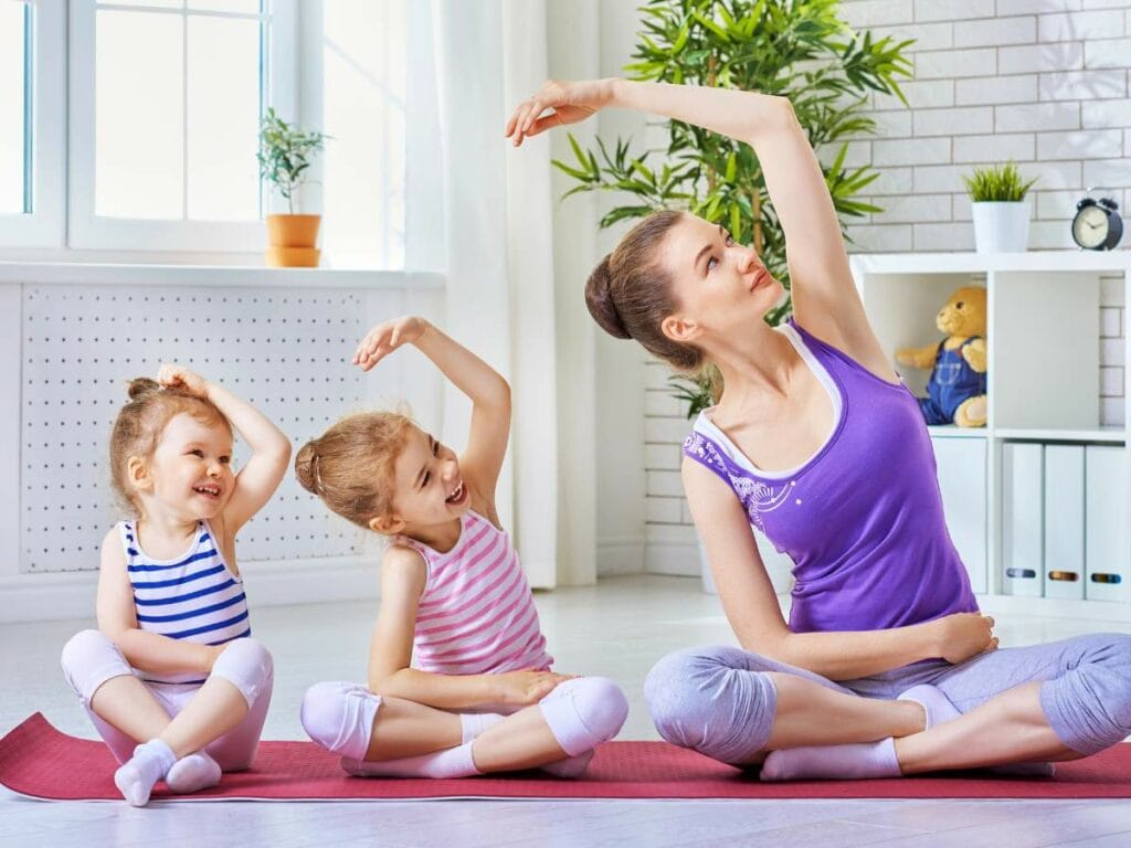 A woman and two children sit cross-legged on yoga mats, stretching their arms overhead.