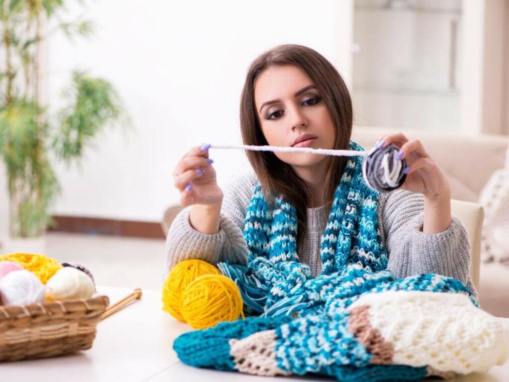 A woman examines yarn while surrounded by knitted items and colorful balls of yarn on a table.