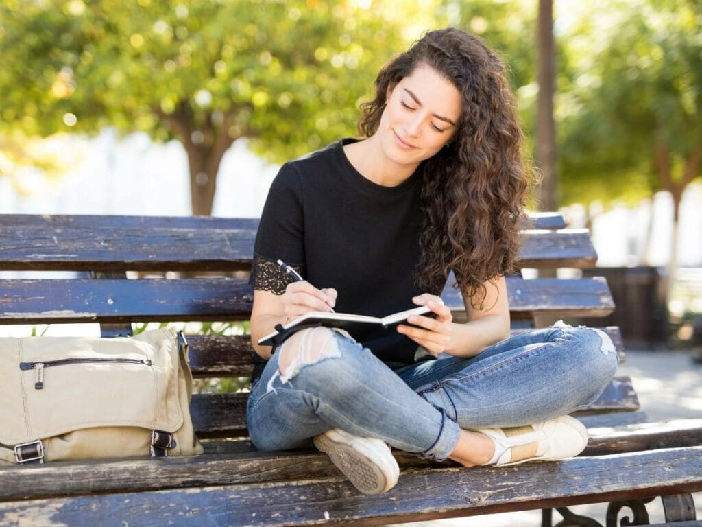 A woman sitting on a park bench is writing in a journal, with a beige bag beside her.