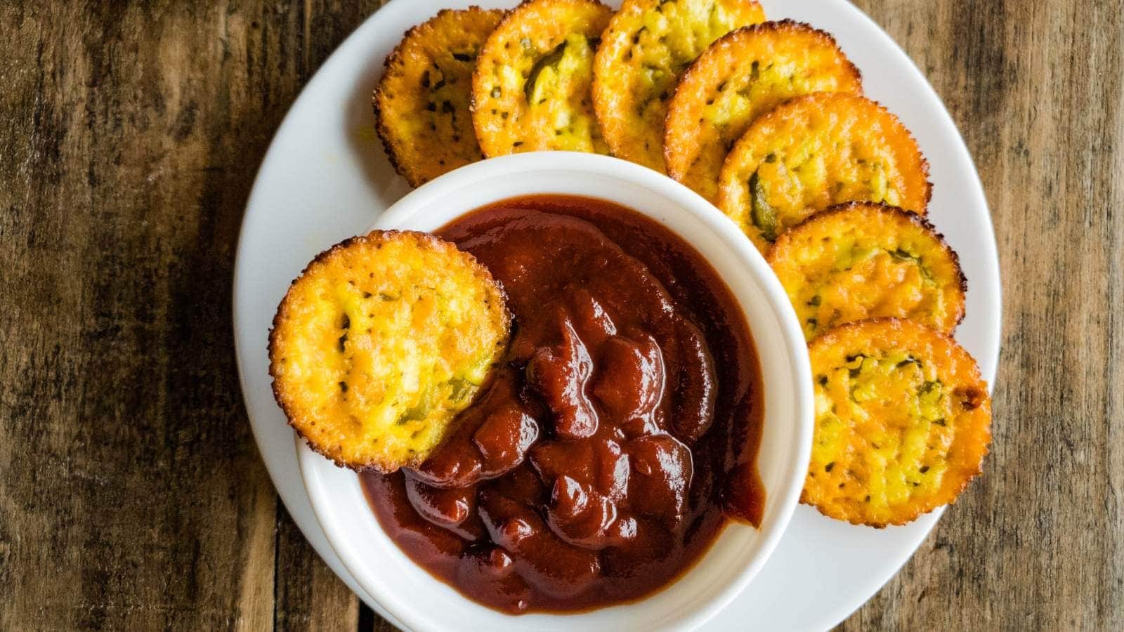 A plate ofpickle chips is arranged around a white bowl filled with thick, dark red sauce on a wooden table.