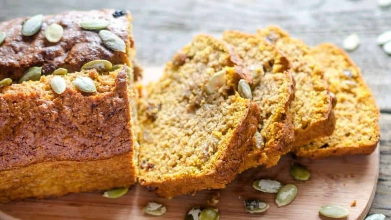 A loaf of pumpkin bread partially sliced on a wooden surface, with visible pumpkin seeds on top and inside the bread slices.