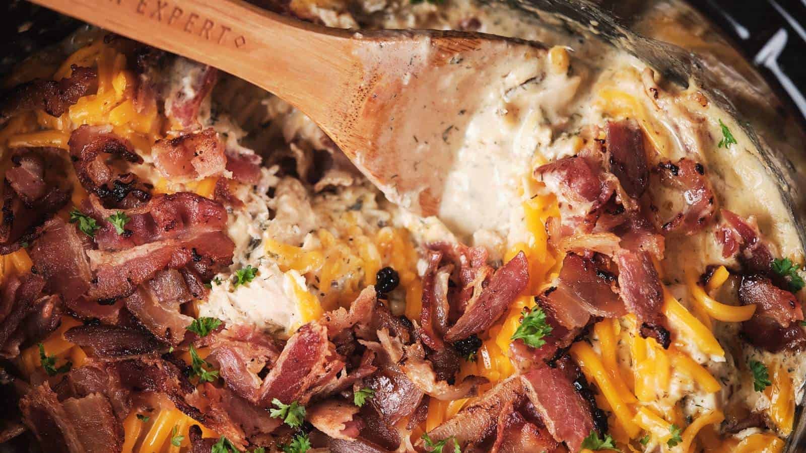 A wooden spoon stirs a dish containing slow cooker crack chicken in a pan.