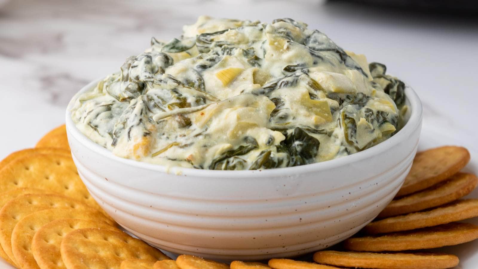 Bowl of spinach artichoke dip surrounded by round crackers on a white surface.