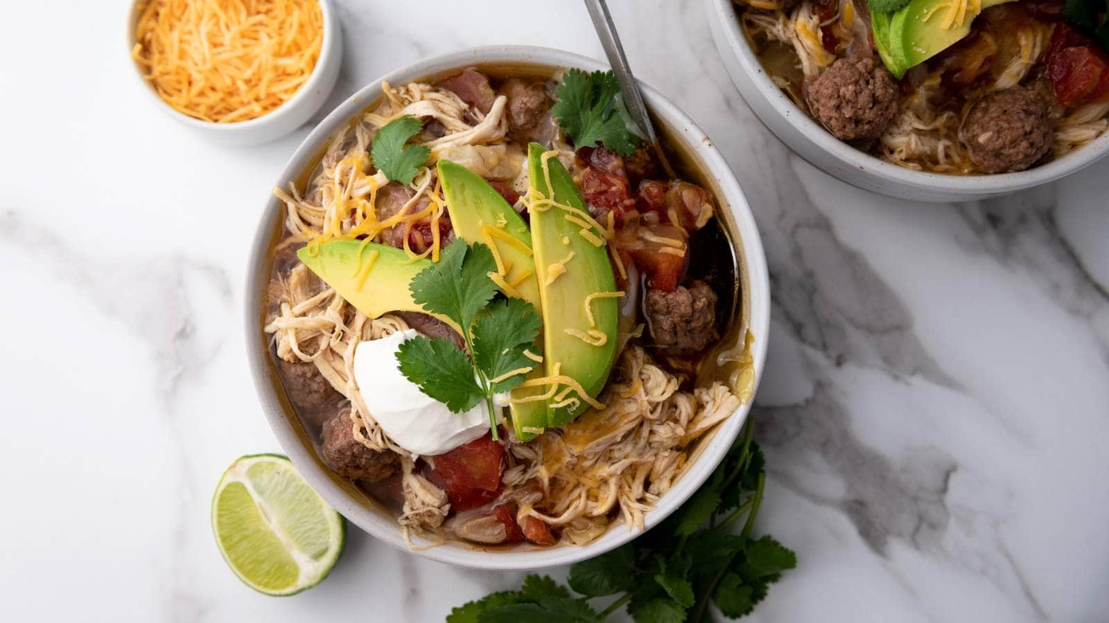 A bowl of chicken and beef soup topped with avocado slices, shredded cheese, sour cream, cilantro, and a lime wedge on the side.