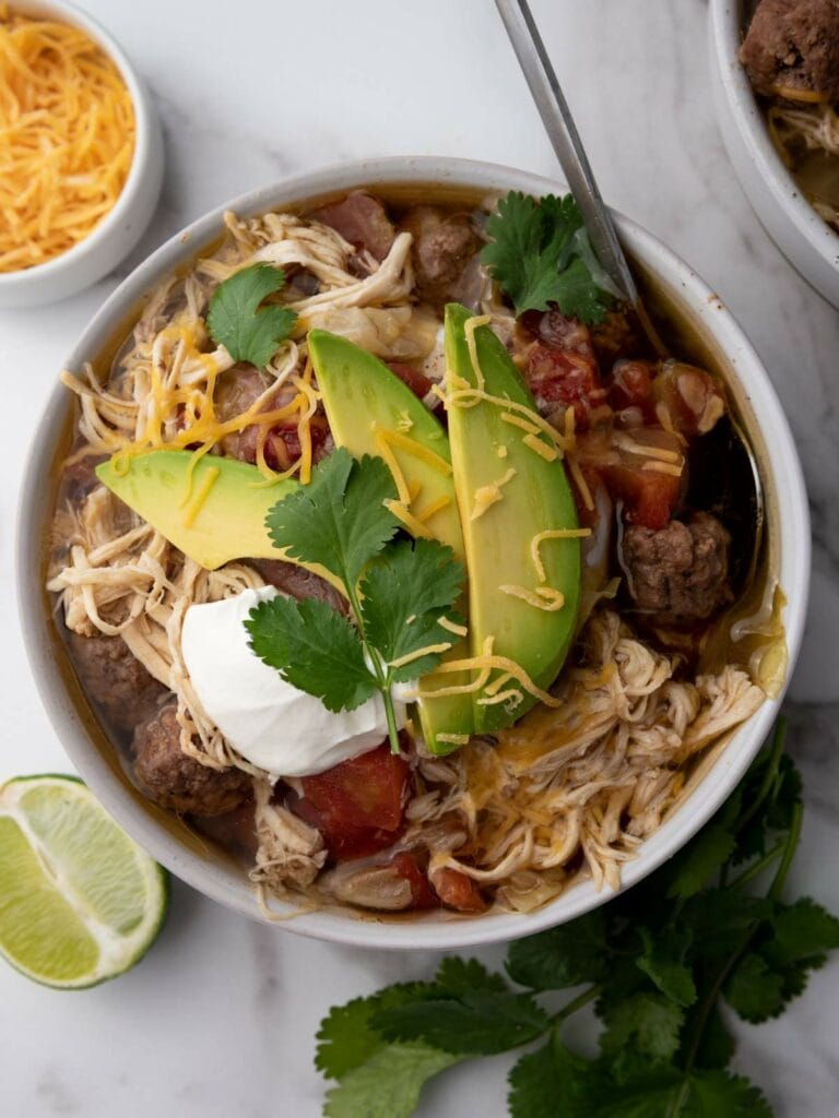 Bowl of taco soup topped with avocado slices, shredded cheese, sour cream, and cilantro.