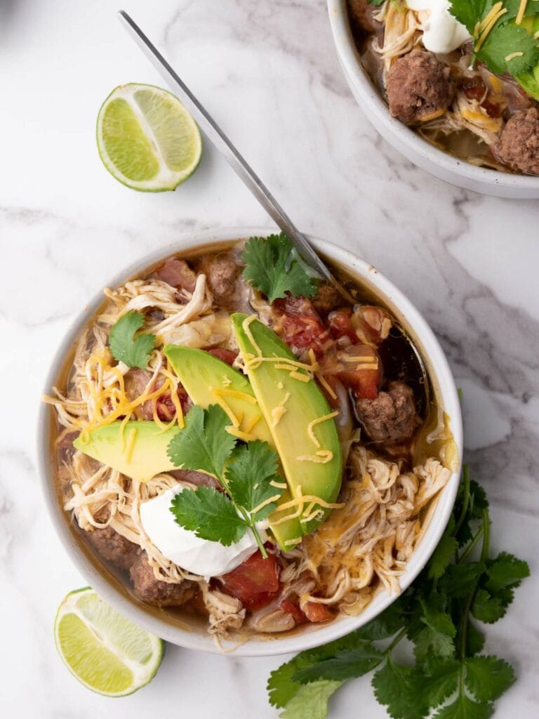 A bowl of taco soup topped with sliced avocado, shredded cheese, sour cream, and cilantro.