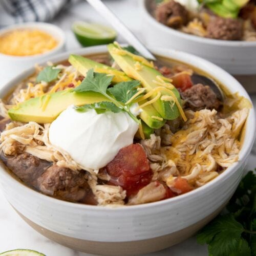 A bowl of taco soup with shredded chicken, avocado slices, sour cream, shredded cheese, and cilantro.