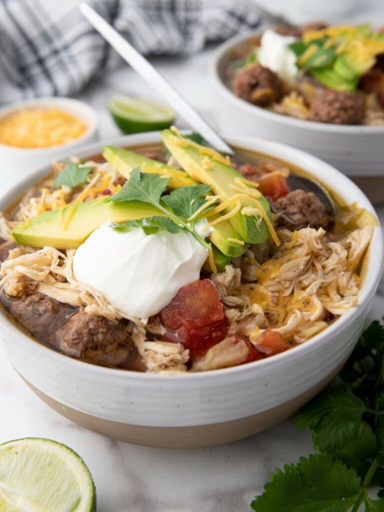 A bowl of taco soup with shredded chicken, avocado slices, sour cream, shredded cheese, and cilantro.