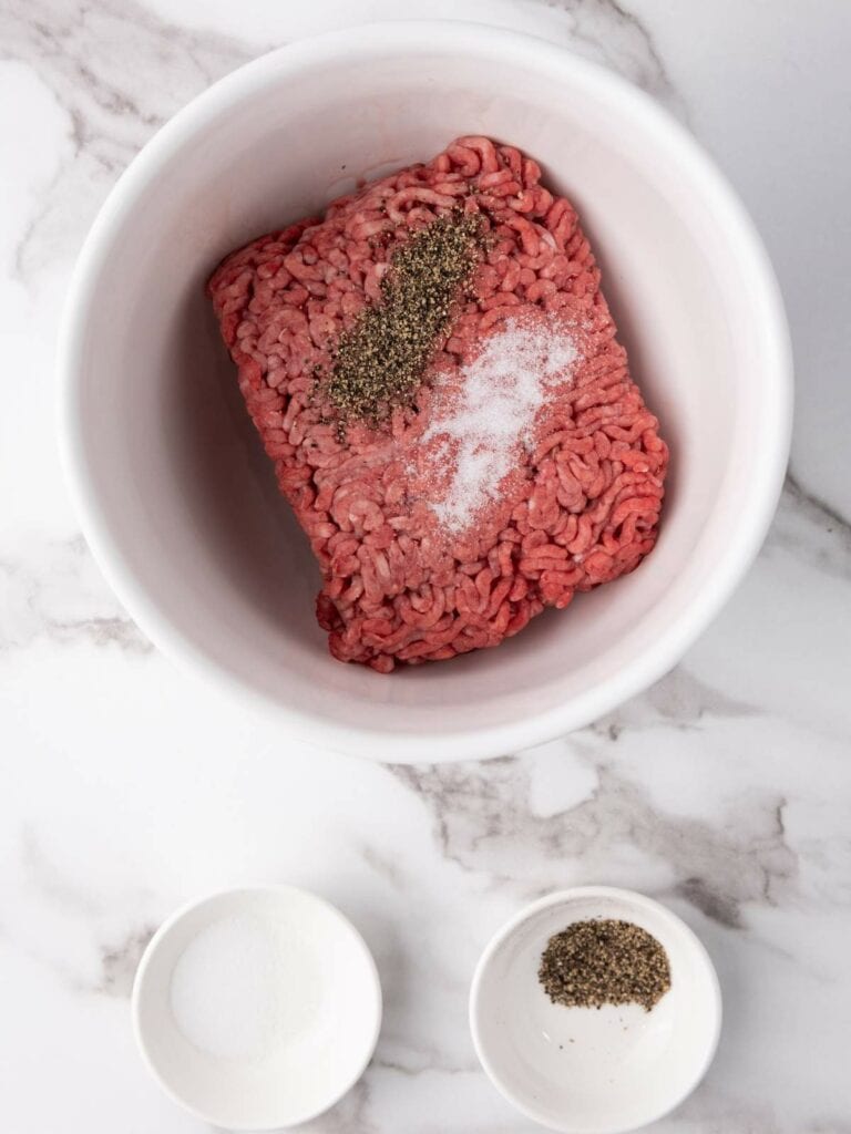 Ground beef with salt and pepper is in a white bowl on a marble surface.