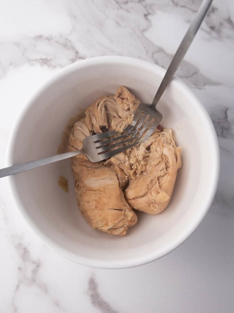 Two forks shredding cooked chicken breast in a white bowl on a marble surface.