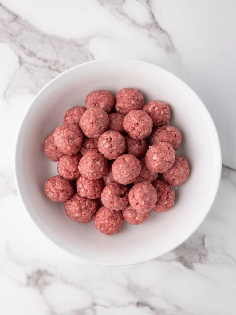 A white bowl filled with raw meatballs on a marble countertop.