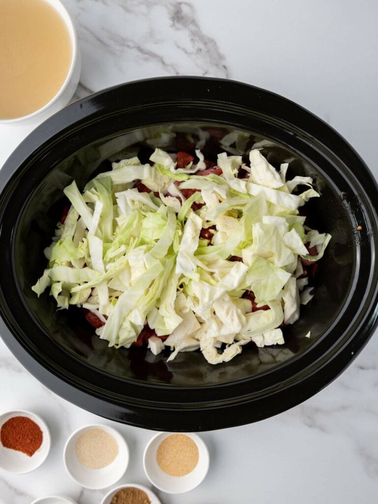 Chopped cabbage in a slow cooker with spices and a bowl of clear broth nearby on a marble surface.