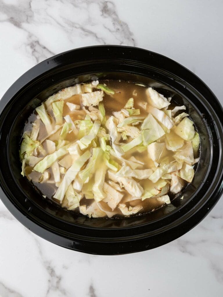 A black crockpot containing pieces of cabbage and chicken in a light broth, placed on a marble surface.