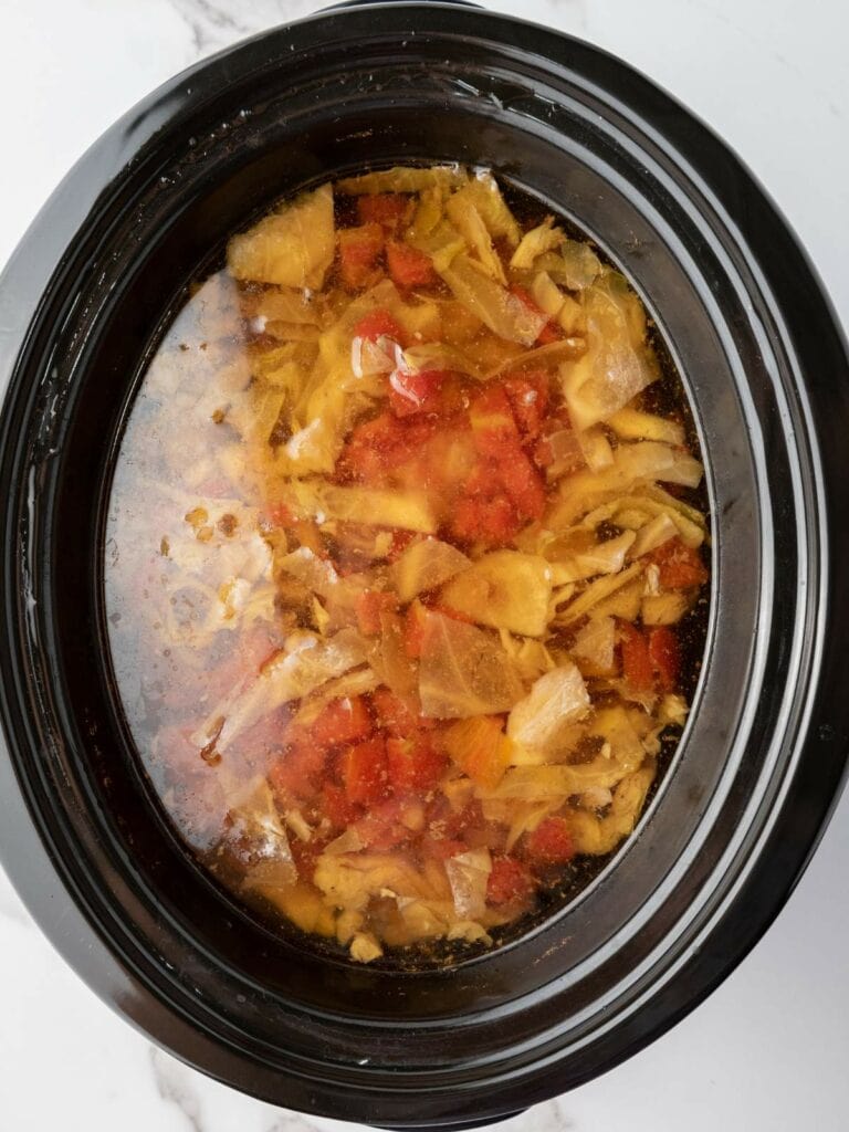 A slow cooker filled with a stew containing chopped cabbage, tomatoes, and broth.