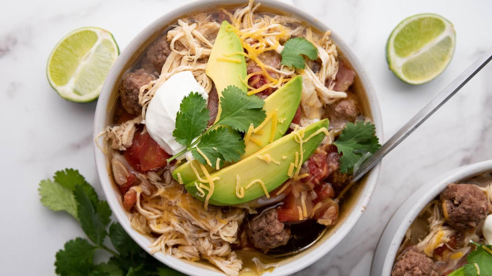 A bowl of taco soup topped with sliced avocado, cilantro, shredded cheese, sour cream.