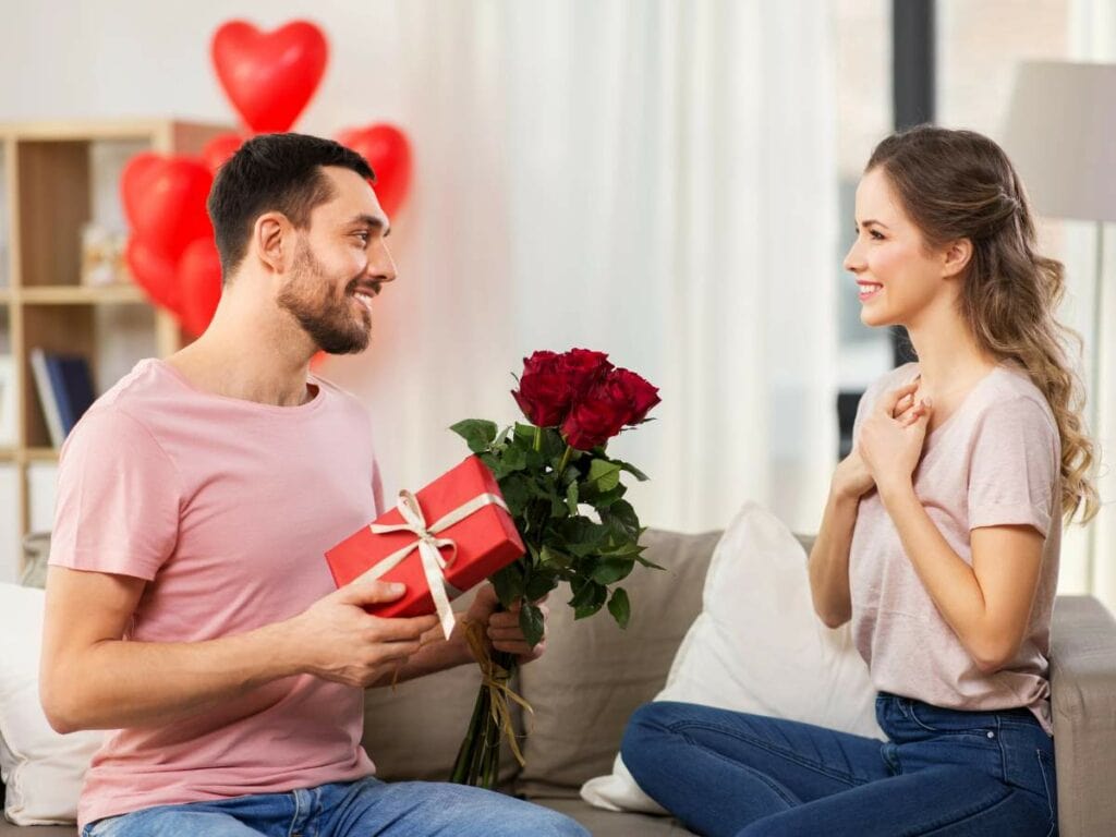 A man on a couch presents a bouquet of red roses and a gift box to a seated woman.