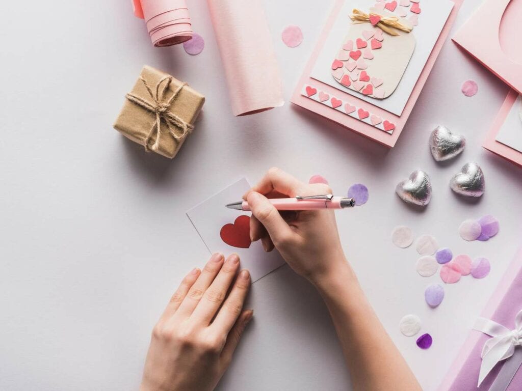 Hands crafting a handmade card with a red heart, surrounded by gift box, pink paper, and heart-shaped decorations.