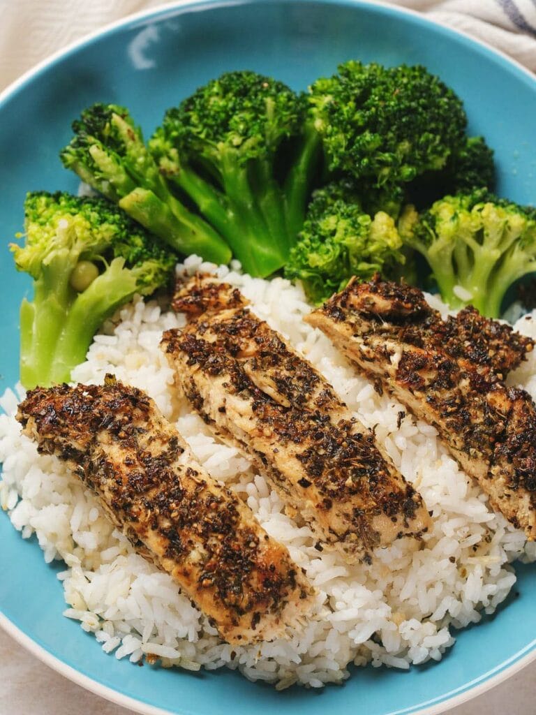 A blue plate with grilled herb-seasoned chicken strips on white rice, accompanied by steamed broccoli.