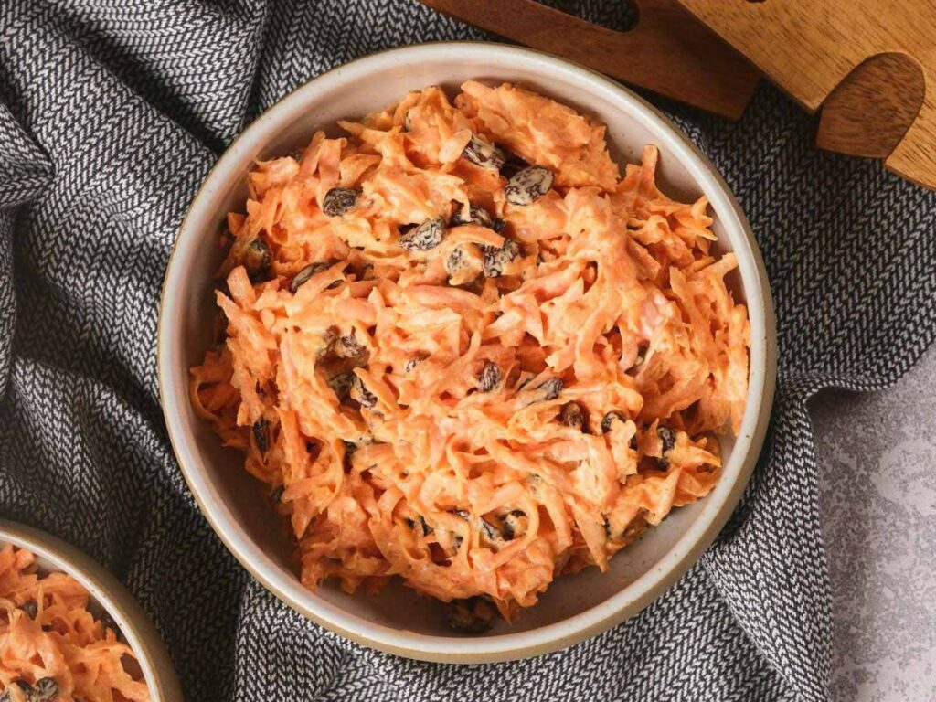 A bowl of carrot raisin salad with creamy dressing on a textured gray cloth.