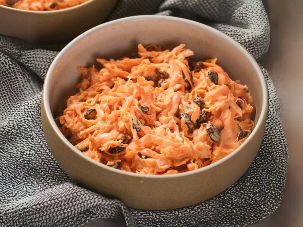 A bowl of carrot salad with raisins on a textured gray cloth.