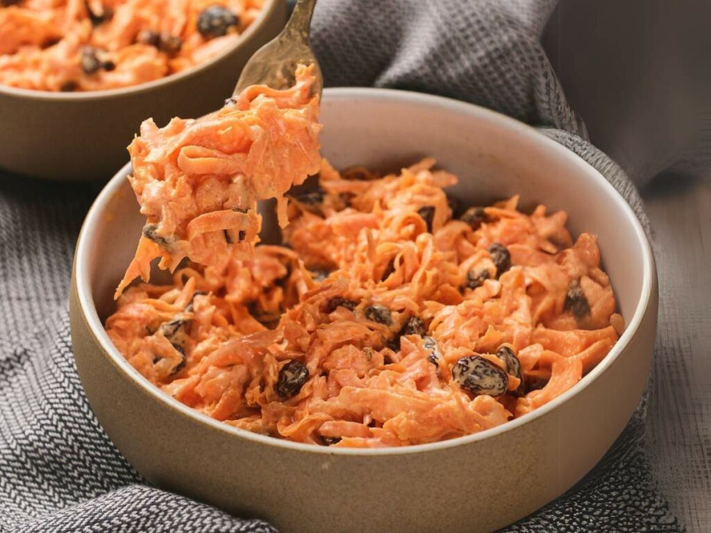 A bowl of carrot salad with raisins and shredded carrots, held with a fork, placed on a dark textured cloth.