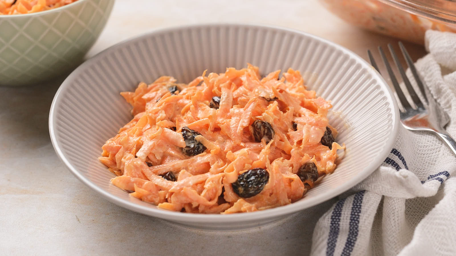 A bowl of carrot raisin salad with a creamy dressing in a white ridged bowl, placed on a light surface.