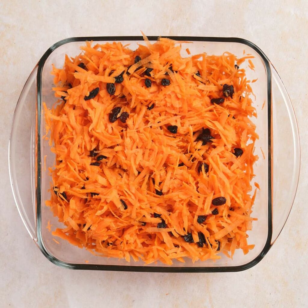 A glass dish filled with shredded carrots and raisins on a light-colored surface.