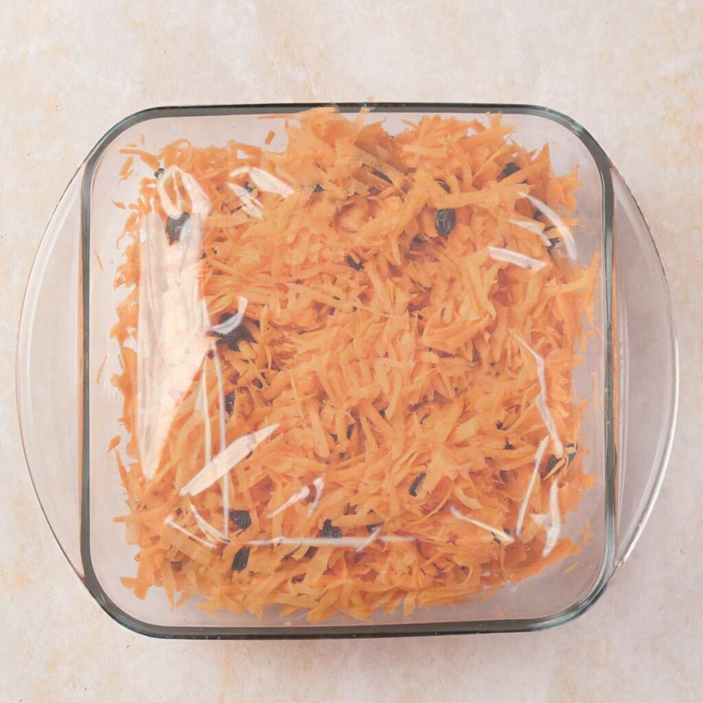 A glass dish filled with shredded carrots and raisins, covered with plastic wrap.