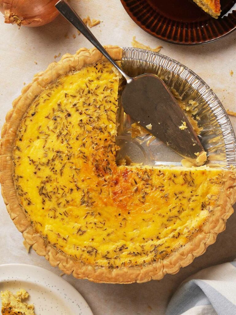 A baked cheese and onion quiche with a slice removed, revealing a creamy filling topped with herbs.