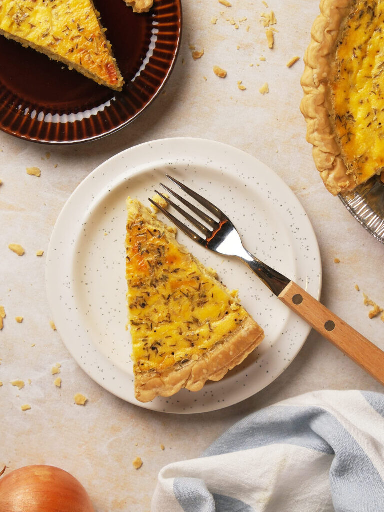 A slice of cheese and onion quiche on a white plate with a fork, surrounded by crumbs.