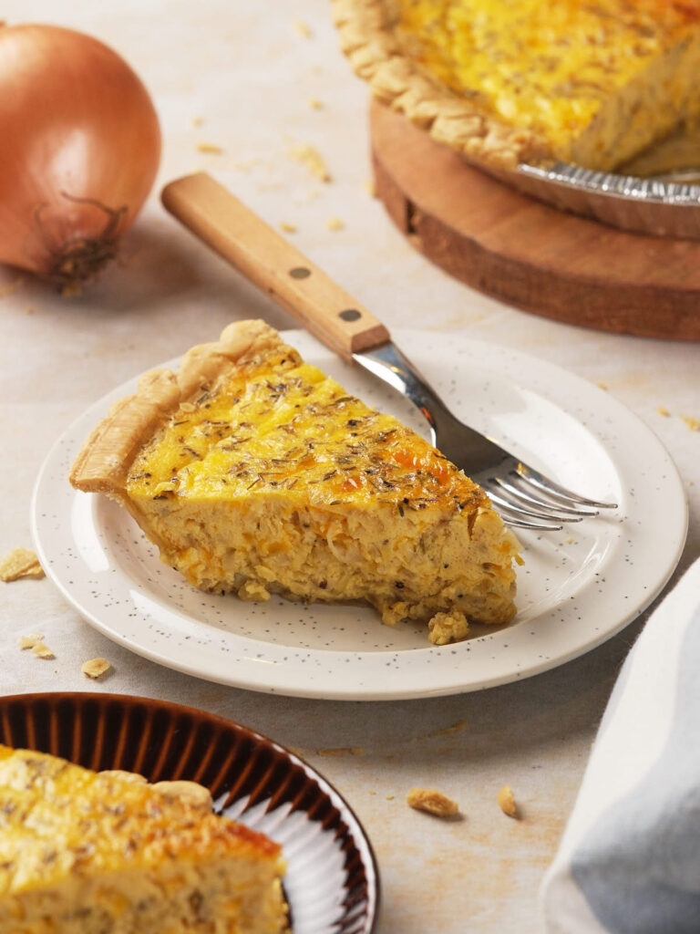 Slice of cheese quiche on a white plate with a fork, set on a light surface.