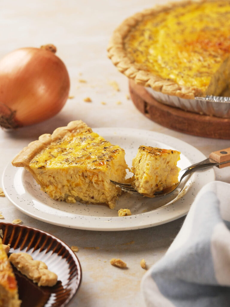 A slice of cheese and onion pie on a plate with a fork. Whole pie and onion in the background.