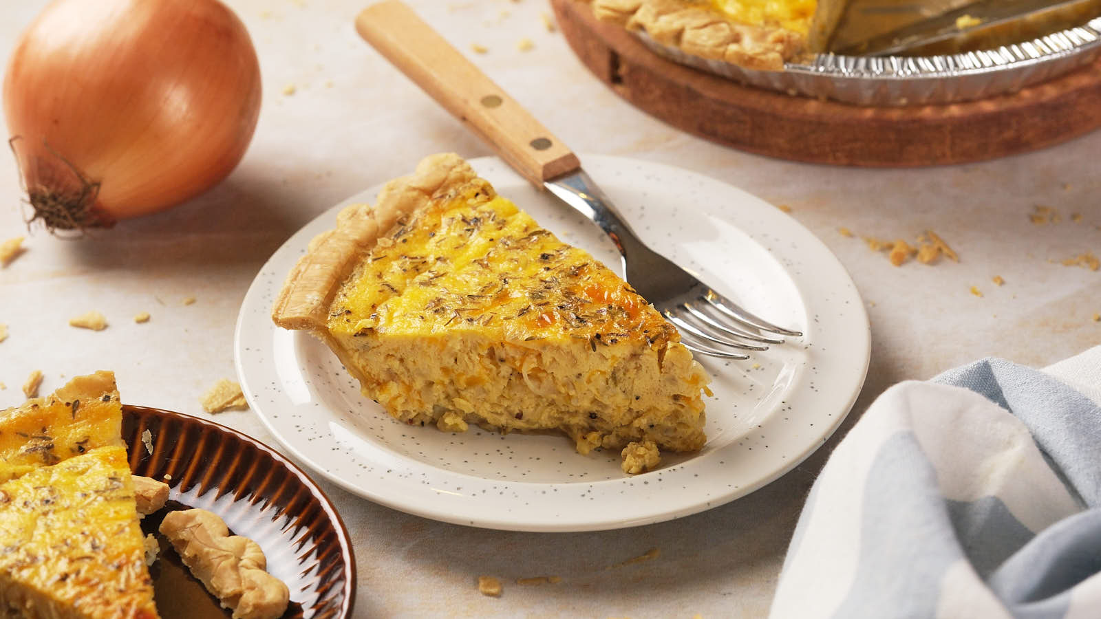 A slice of cheese and onion quiche on a white plate with a fork, next to a whole quiche in a tin.