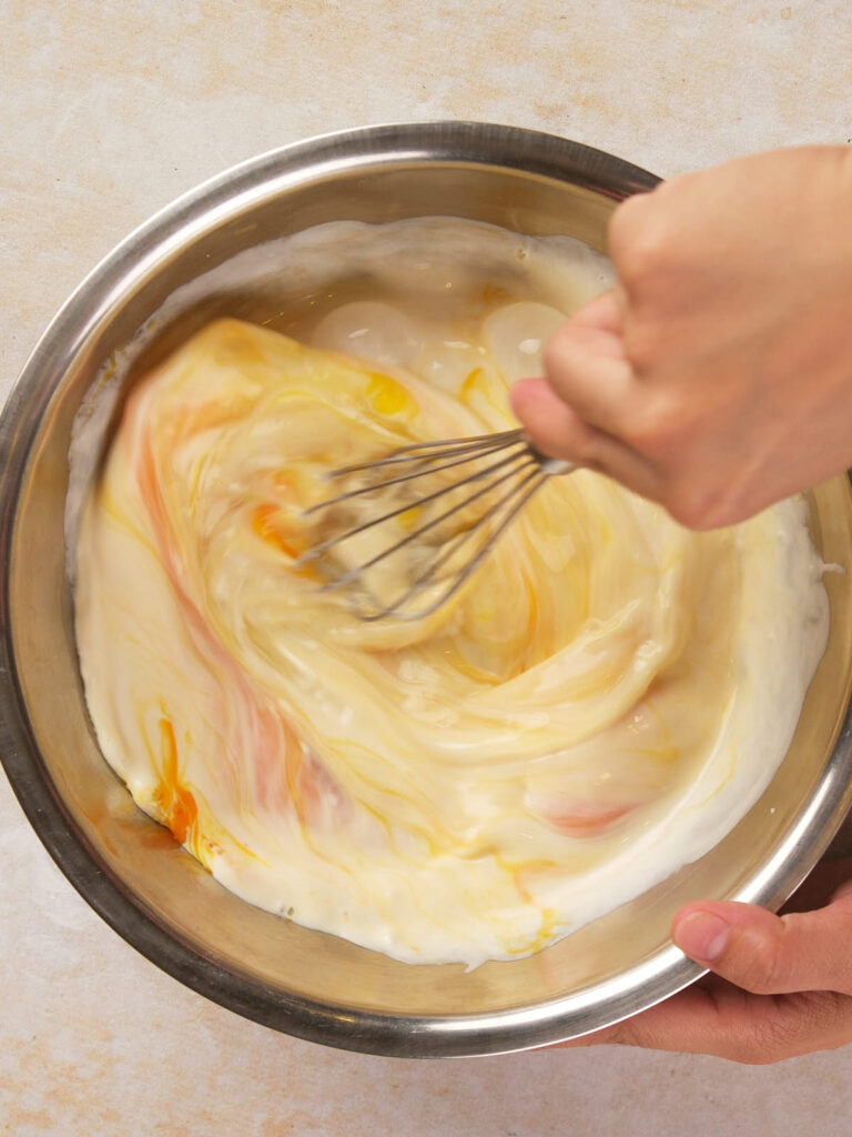 A person is whisking cream with orange food coloring in a metal bowl.