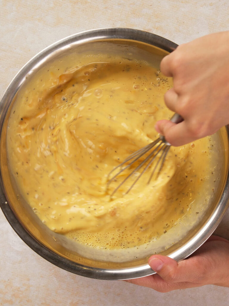 A person whisking a yellow mixture with visible herbs in a stainless steel bowl.