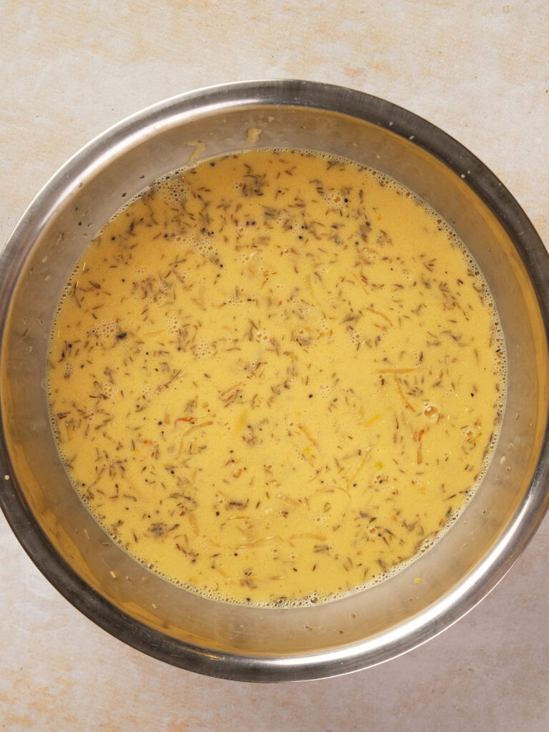 A metal bowl containing a yellow liquid mixture with visible herbs and spices.