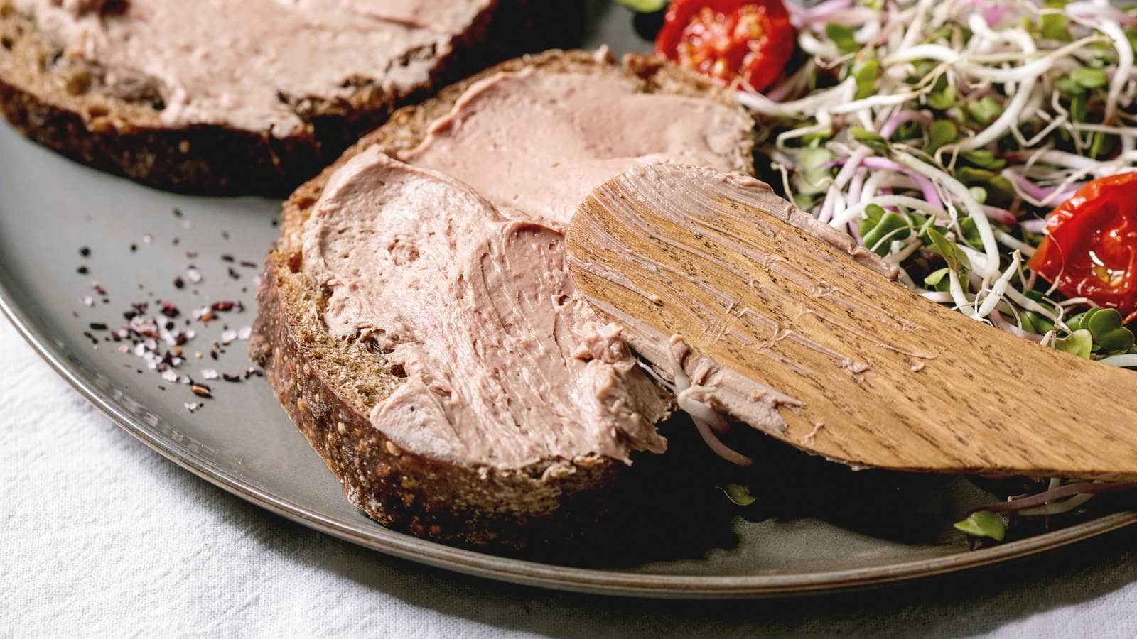 Two slices of bread with chicken liver spread on a grey plate, with a wooden knife resting on one slice.