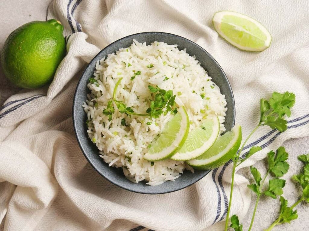 Bowl of chipotle rice garnished with lime wedges and cilantro on a cloth background.