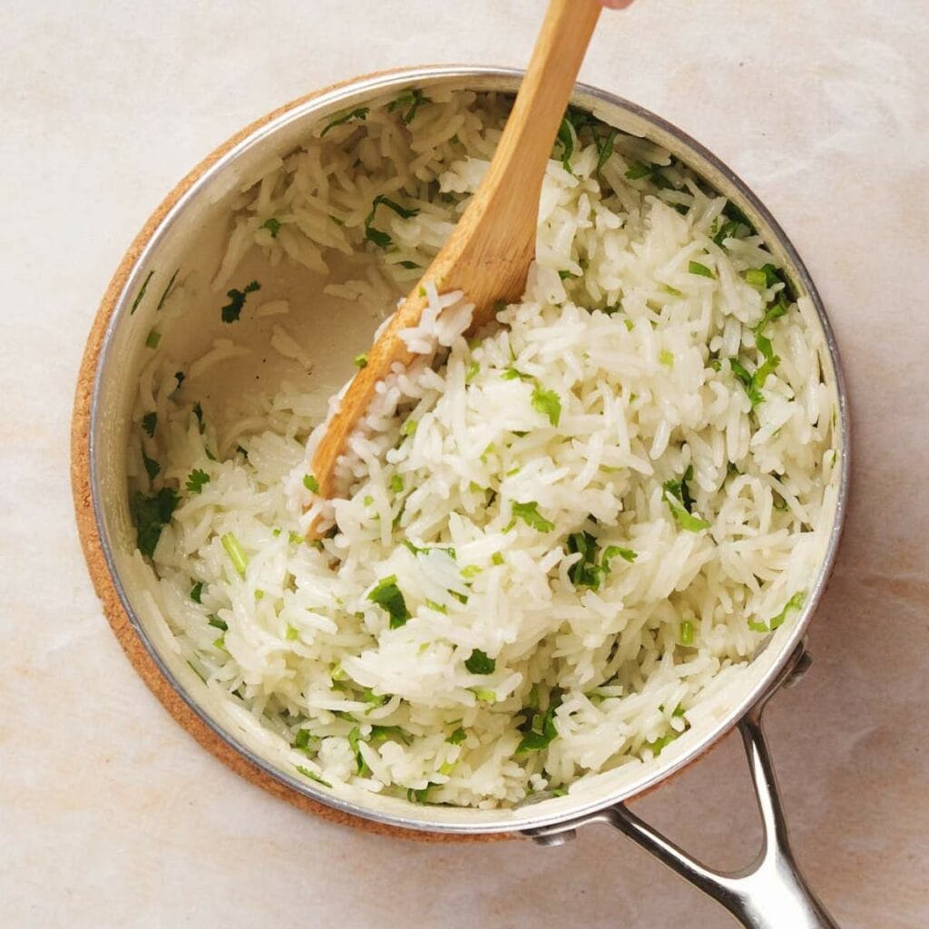 A pot of cooked rice mixed with chopped herbs, being stirred with a wooden spoon.