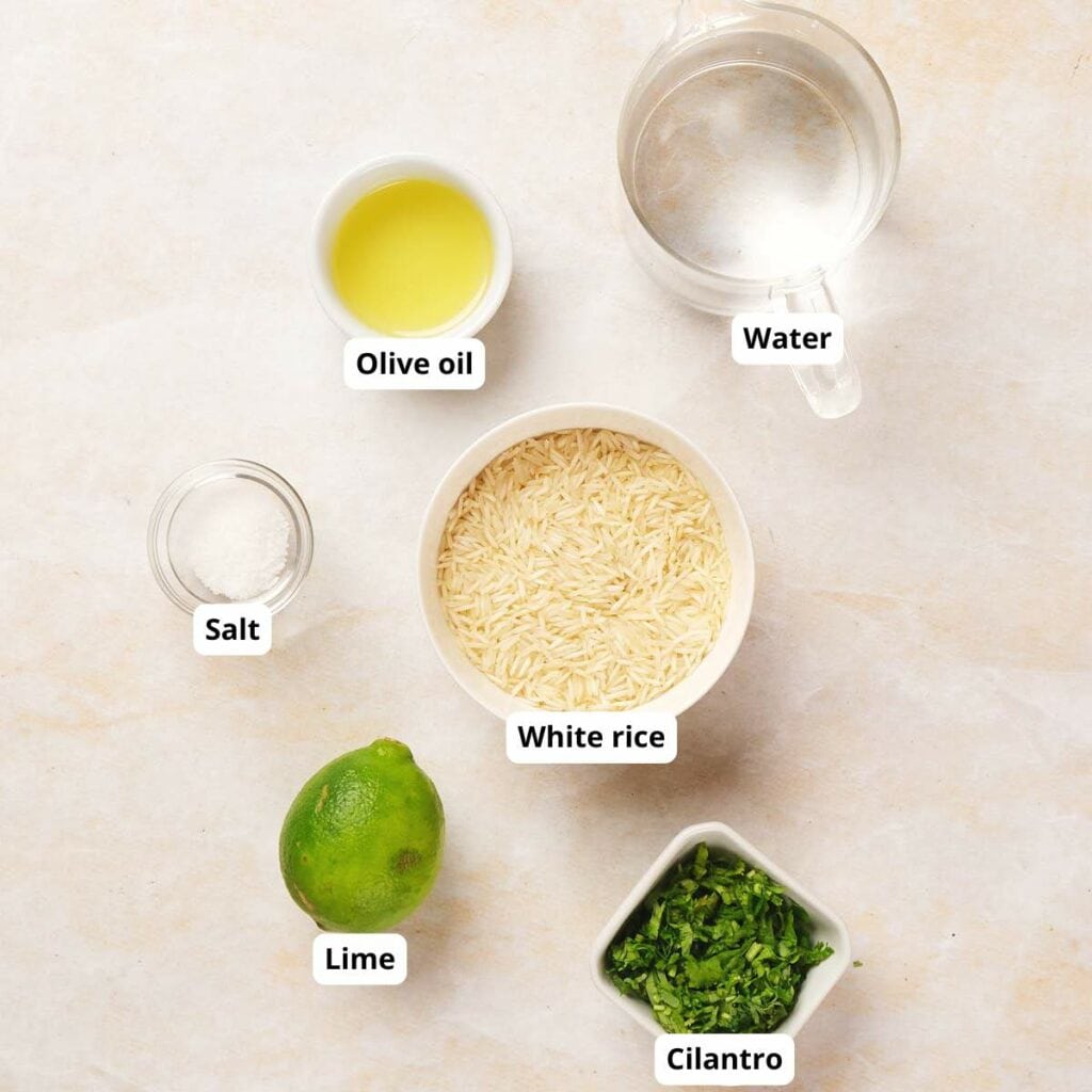 White rice, olive oil, water, salt, a lime, and cilantro are arranged on a light surface.