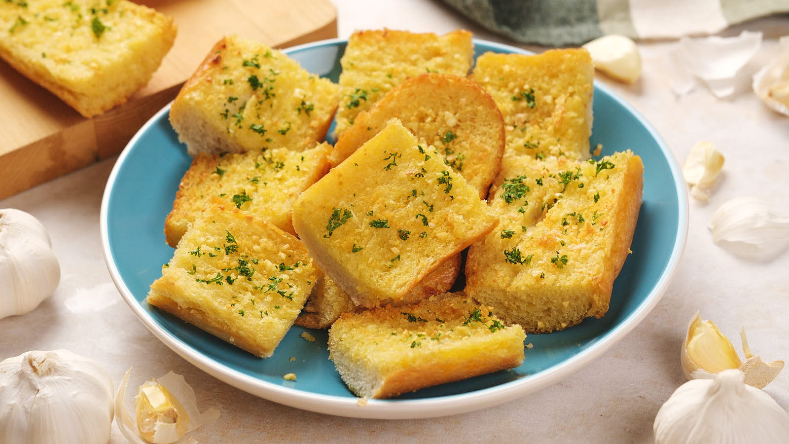 Slices of garlic bread topped with parsley on a blue plate, surrounded by garlic cloves.