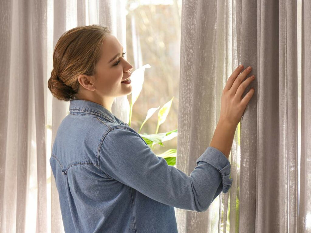 A woman in a denim shirt stands by a window, touching grey curtains, with sunlight filtering through.