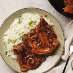 A plate with two glazed pork chops and a serving of white rice garnished with parsley. A fork and a napkin are beside the plate.