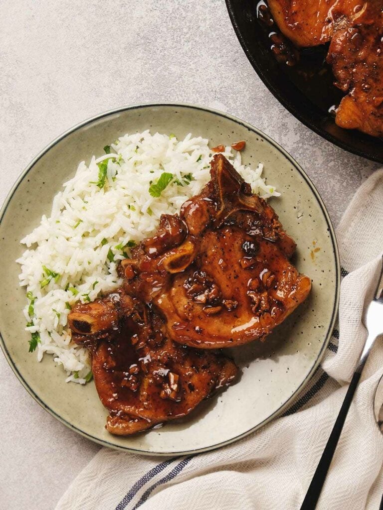 A plate with two glazed pork chops and a serving of white rice garnished with parsley. A fork and a napkin are beside the plate.