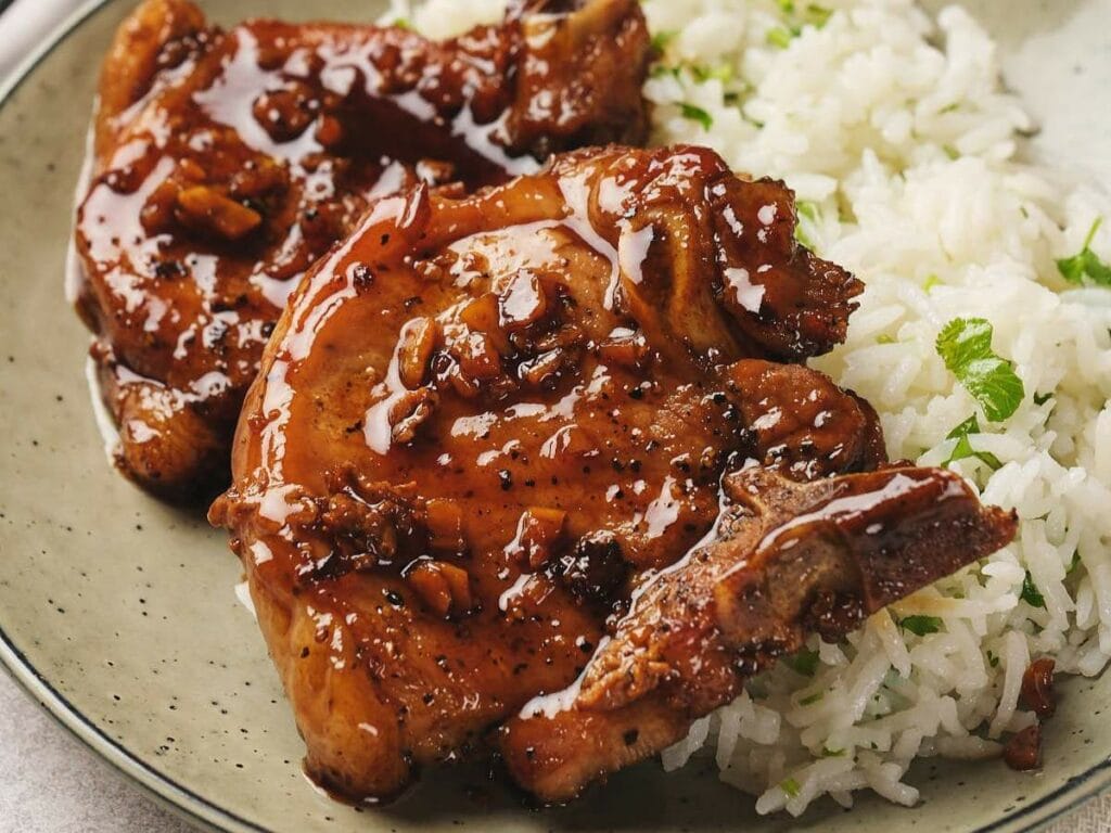 Plate with glazed pork chops seasoned with pepper and garlic, served alongside white rice with green herbs.