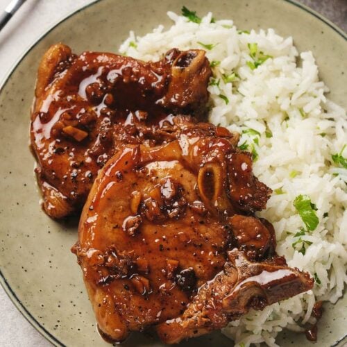 Two glazed pork chops with a glossy sauce sit on a plate beside fluffy white rice garnished with parsley.