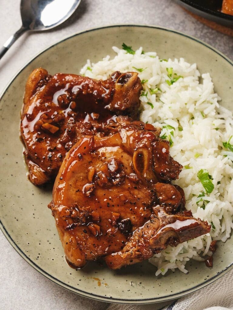 Two glazed pork chops with a glossy sauce sit on a plate beside fluffy white rice garnished with parsley.